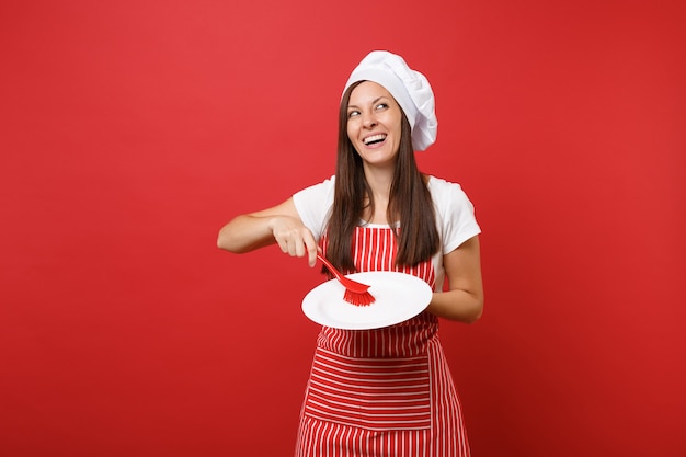 Huisvrouw vrouwelijke chef-kok of bakker in gestreepte schort, wit t-shirt, toque chef-koks hoed geïsoleerd op rode muur achtergrond. Vrouw houdt witte plaat met borstel voor het afwassen. Bespotten kopie ruimte concept.