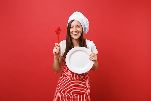 Huisvrouw vrouwelijke chef-kok of bakker in gestreepte schort, wit t-shirt, toque chef-koks hoed geïsoleerd op rode muur achtergrond. Vrouw houdt witte plaat met borstel voor het afwassen. Bespotten kopie ruimte concept.