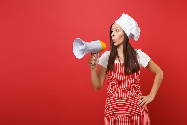 Huisvrouw vrouwelijke chef-kok of bakker in gestreepte schort, wit t-shirt, toque chef-koks hoed geïsoleerd op rode muur achtergrond. vrouw schreeuwt in megafoon, kondigt kortingenverkoop aan. mock-up kopie ruimteconcept.