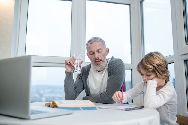 Huiswerk. Man met grijze baard zit met zijn zoon aan tafel terwijl hij les geeft