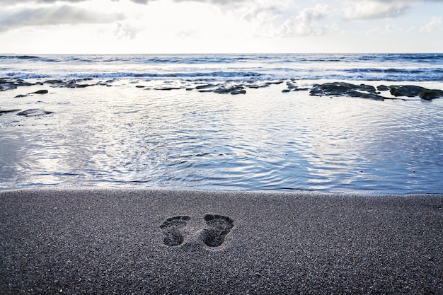 Human barefoot footprints in sand heading to sea awaiting incoming wave sunny cloudy sunrise active healthy living and personal growth concept copy space