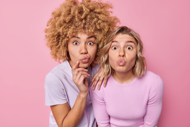 Photo human facial expressions concept surprised young beautiful women stare at camera keep lips folded react on amazing news dressed in casual clothes isolated over pink background admire something