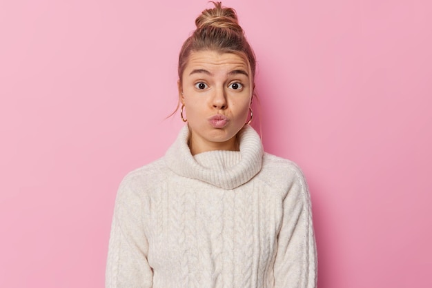 Human facial expressions concept Surprised young cute European woman keeps lips folded looks wondered has fair hair gathered in bun wears white jumper with collar isolated over pink background