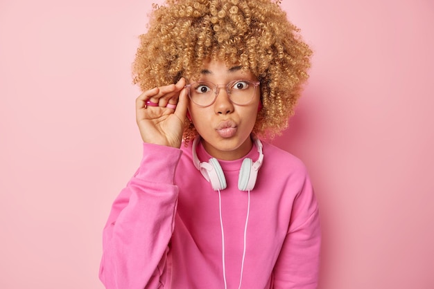 Human facial expressions lovely surprised curly haired woman\
keeps lips rounded wears transparent eyeglasses and casual jumper\
uses headphones for listening music poses indoor against pink\
wall
