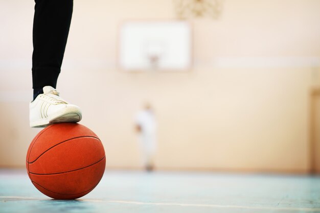 コンクリートの床のバスケットボールに人間の足が休む。木の床にある1つのバスケットボールとスニーカーの写真。