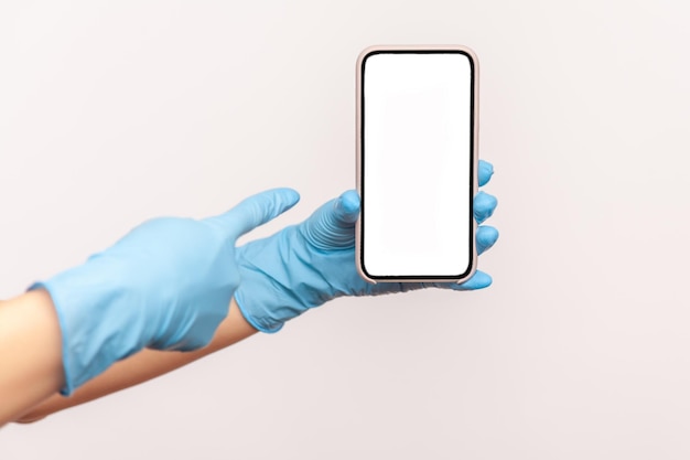 Human hand in blue surgical gloves holding and showing smart phone and pointing at empty display.