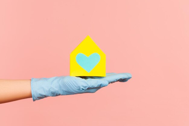 Human hand in blue surgical gloves holding yellow paper house exterior in hand with love and care.