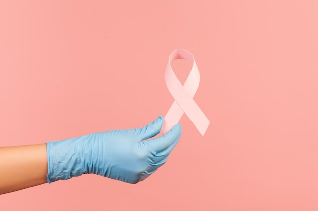 Human hand in blue surgical gloves showing and holding pink ribbon, symbol of breast cancer.