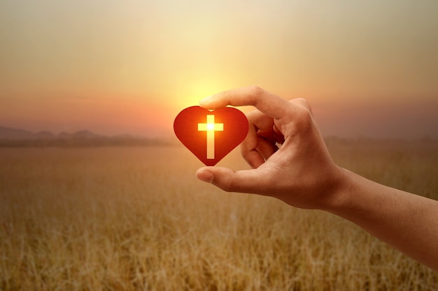 Human hand holding a red heart with a Christian cross with a sunrise sky