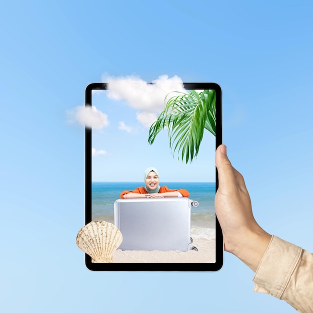 A human hand holding tablet with a screen view of an Asian girl in a scarf sitting with a suitcase on the beach with an ocean view and blue sky