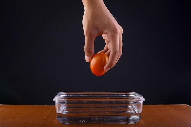 A human hand holding a tomato over a glass container filled with water.