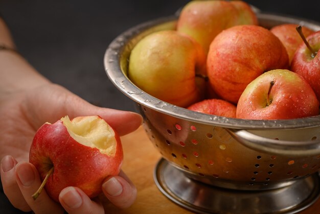 Human hand holds bitten apple taken from metallic bowl full of red ripe apples. Healthy eating. Apple pie ingridients. Cooking at home