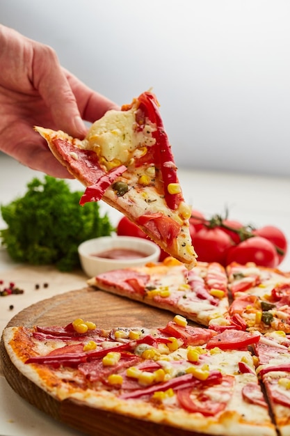 The human hand takes Freshly baked tasty pepperoni pizza with salami mozzarella cheese corn and pepper served on wooden background with tomatoes sauce and herbs Food delivery concept Restaurant menu