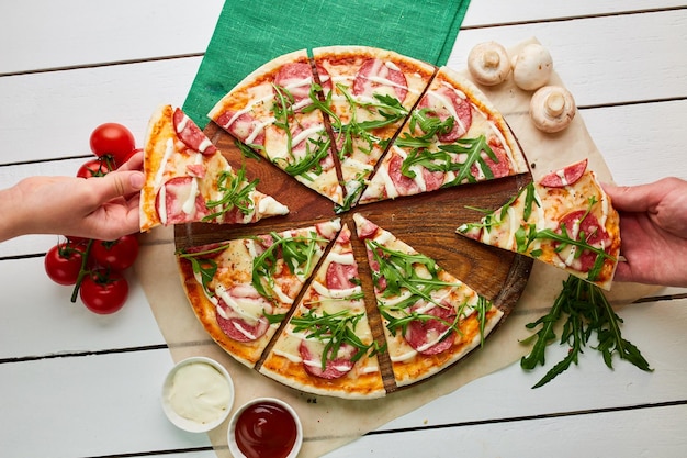 The human hand takes freshly baked tasty pepperoni pizza with salami mozzarella cheese and rukkola served on wooden background with tomatoes sauce and herbs Food delivery concept