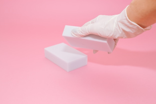 Human hand in white glove holds a white melamine household sponge for cleaning on pink background. 