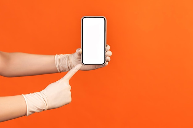 Human hand in white surgical gloves holding and showing smart phone and pointing at empty display