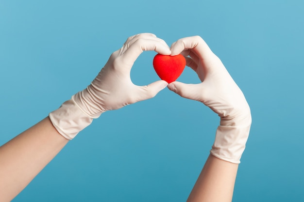 Human hand in white surgical gloves holding smal red heart shape in hand.