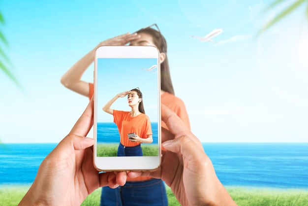 Human hand with the mobile phone taking a picture of Asian woman traveling on the field with seascape background