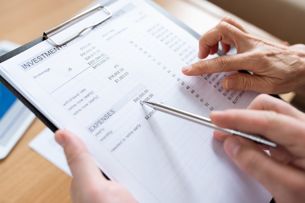 Human hand with pen pointing at sum of expenses while discussing financial paper with another person