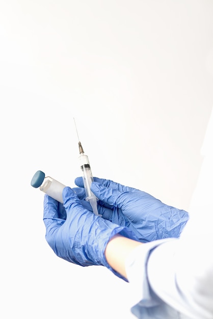 Human hands in blue protective gloves hold syringe and ampoule with covid-19 vaccine on light