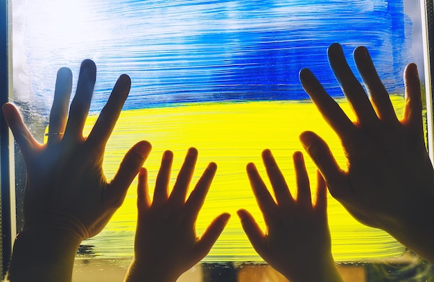 Человеческие руки родителя и ребенка касаются картины желто-голубого флага Украины на окне