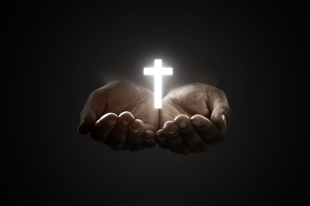 Human hands praying to god with shiny christian cross over black background