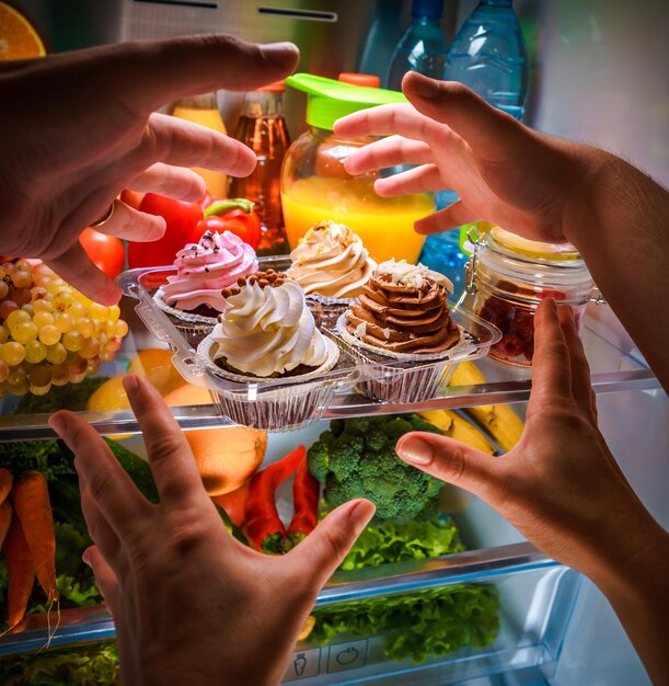 Human hands reaching for sweet cake at night in the open refrigerator