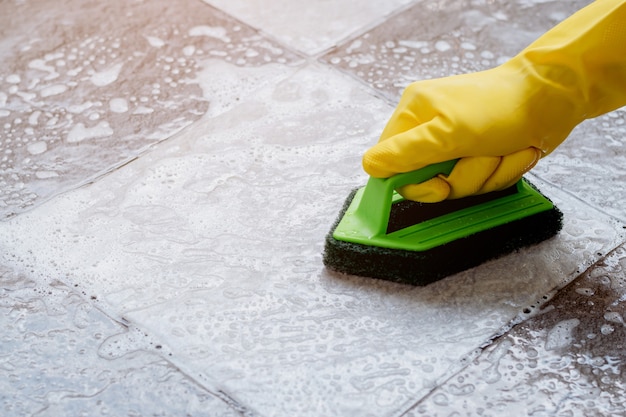 写真 黄色のゴム手袋を着用している人間の手は、緑色のプラスチック製フロアスクラバーを使用して、床掃除機でタイルの床をこすります。