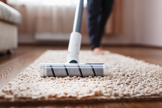 Human legs and a white turbo brush of a cordless vacuum cleaner cleans the carpet in the house