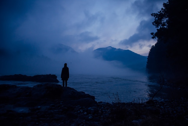 Human silhouette in a blue haze on the background of mountains and river. Thick fog in the evening twilight. Mysterious atmosphere. Concept of solitude, reflection, meditation.