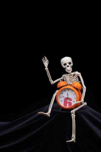 A human skeleton waving his hand greeting holding an orange alarm clock on a black background vertical frame