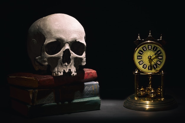 Cranio umano su vecchi libri vicino al retro orologio vintage su sfondo nero sotto il fascio di luce.