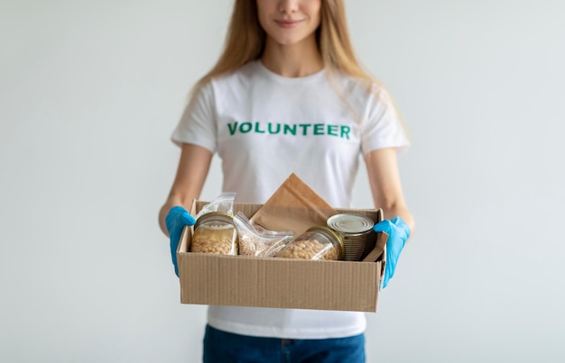 Фото Концепция гуманитарной помощи молодая женщина в волонтерской футболке и медицинских перчатках держит коробку для пожертвований еды