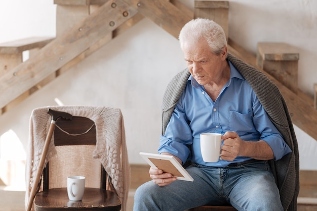 Humeurige ongelukkige senior man kijkt naar de foto van zijn vrouw en mist haar terwijl ze thee drinkt