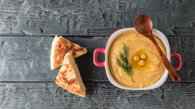 Hummus in een paarse kom met een lepel en twee sneetjes donker brood op tafel