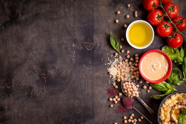 Ingredienti hummus. ceci, tahini, olio d'oliva, semi di sesamo, sommacco, erbe aromatiche su fondo di legno rustico scuro.