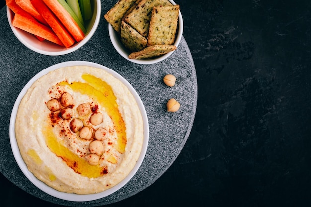 Hummus met olijfolie en kikkererwten in kom met groentesticks en knapperig brood op donkere stenen achtergrond Bovenaanzicht met kopieerruimte