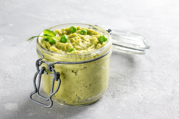 Hummus van avocado in een glazen pot met kikkererwten, groene erwten, kruiden op grijze achtergrond