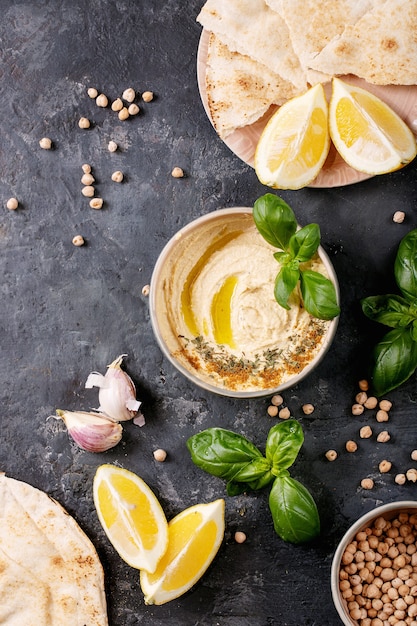 Hummus with olive oil, pita bread and ground cumin in ceramic bowl served with lemons, basil and chickpeas over dark texture surface. Top view, flat lay