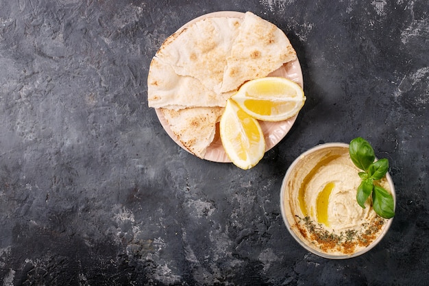 Hummus with olive oil, pita bread and ground cumin in ceramic bowl served with lemons, basil and chickpeas over dark texture surface. Top view, flat lay