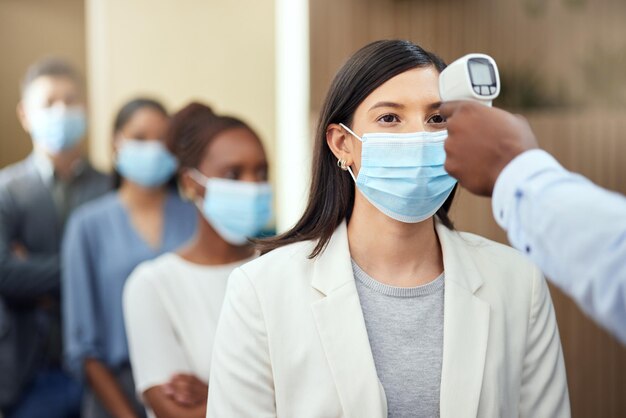 Foto hun temperatuur laten opnemen bijgesneden opname van een aantrekkelijke jonge zakenvrouw die een masker draagt en haar temperatuur laat opnemen terwijl ze vooraan in een rij in haar kantoor staat