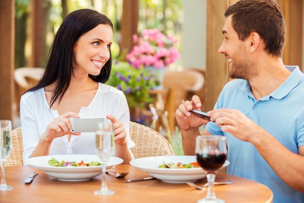 Hun tijd samen vastleggen. Gelukkig jong liefdevol stel dat foto's maakt van hun eten en glimlacht terwijl ze samen in het openluchtrestaurant ontspannen