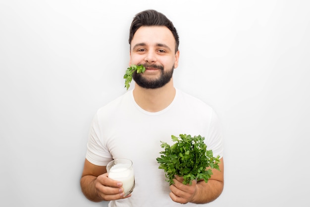 Hungru and funny young man stands and poses. He look at camera. Guy hold glass of kefir and bunch of parsley in hands. Also there are some part of green plant in his mouth. Isolated on white