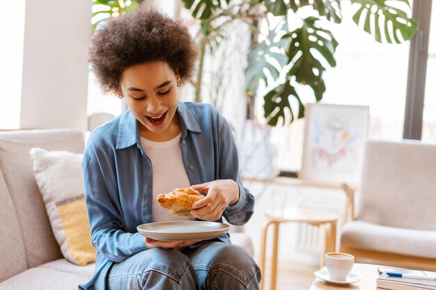 Hungry African American woman eating tasty croissant while sitting on comfortable sofa in cafe