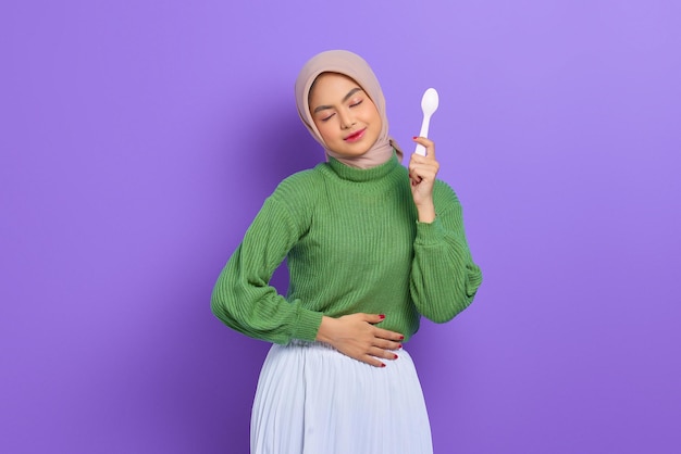 Hungry beautiful Asian woman in green sweater and hijab holding a spoon and think of tasty food isolated over purple background