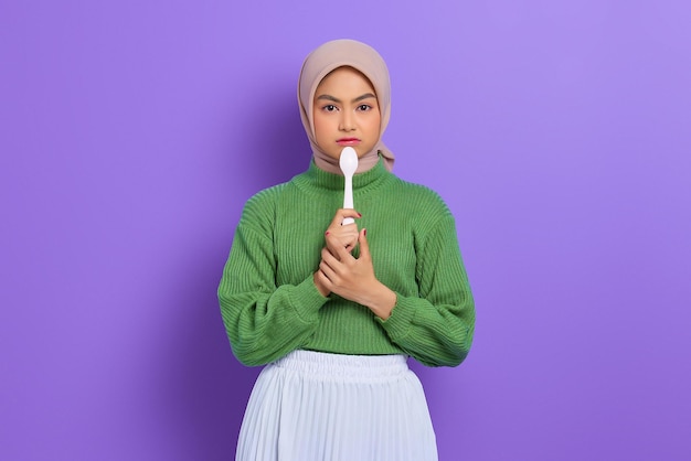 Hungry beautiful Asian woman in a green sweater holding a spoon in her mouth dream of a tasty meal isolated over purple background