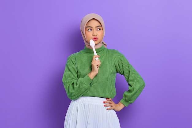 Hungry beautiful asian woman in green sweater holding a spoon\
in mouth think of tasty food isolated over purple background