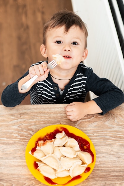 写真 灰色のジャケットを着てテーブルに座って、キッチンで餃子を食べる空腹の子供