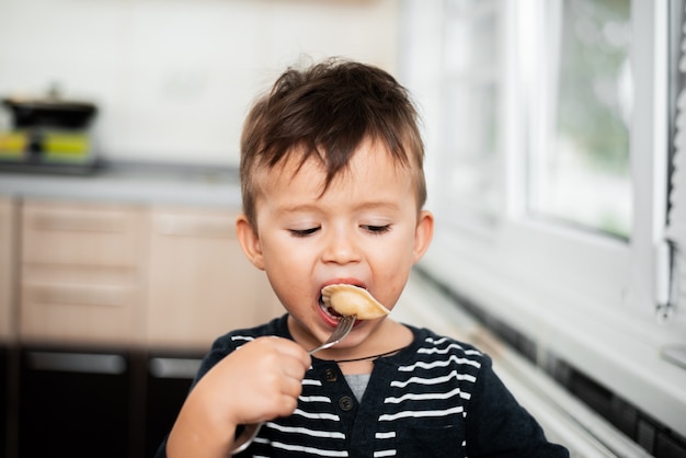 Bambino affamato che mangia gnocchi in cucina, seduto al tavolo con una giacca grigia