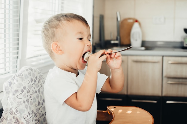 A hungry child is eating dumplings in the kitchen sitting in a chair in a white t-shirt